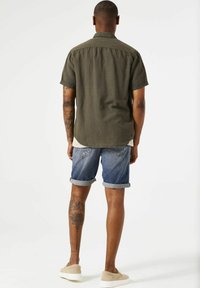 Camisa de oliva de manga corta con dobladillo recto, combinada con pantalones cortos de mezclilla enrollados. Zapatos de color beige claro, tatuajes visibles en brazos y piernas.