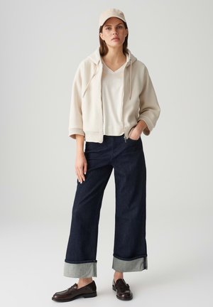 Beige hooded sweatshirt, white v-neck tee, dark denim wide-leg pants with light cuff, and brown loafers, topped with a beige cap.