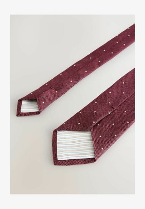 Cravatta bordeaux con un tessuto texturizzato, caratterizzata da piccoli punti bianchi e una fodera interna a righe. Le estremità sono appuntite e leggermente più larghe.