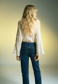 Blusa bianca testurizzata con maniche lunghe e jeans in denim blu, vista da dietro su uno sfondo sfumato.