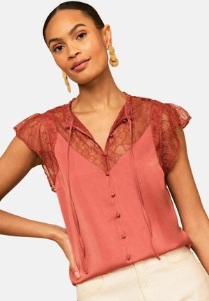 Femeie purtând un top fără mâneci, de culoare ruginiu, cu inserție de dantelă și nasturi în față, asortat cu pantaloni crem și cercei cu discuri aurii.