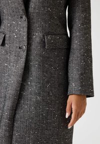 Cappotto a motivo a zig zag in bianco e nero, caratterizzato da un tessuto testurizzato, revers a punta e tasche laterali. La manica è leggermente arrotolata.