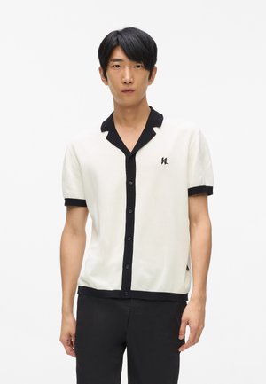 Kortærmet striktrøje i hvid med sort krave og knaplukning, med et logo på brystet og kontrasterende manchetter.