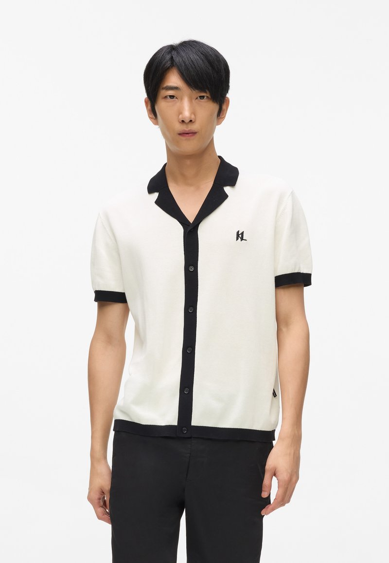 Rövid ujjú kötött póló fehér színben, fekete gallérral és gombos állal, a mellkason logóval és kontrasztos mandzsettákkal.