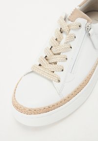 Sneaker en cuir blanc avec des lacets tressés beige et une semelle en jute tressé. Présente une fermeture éclair et un accent en daim marron sur le talon.