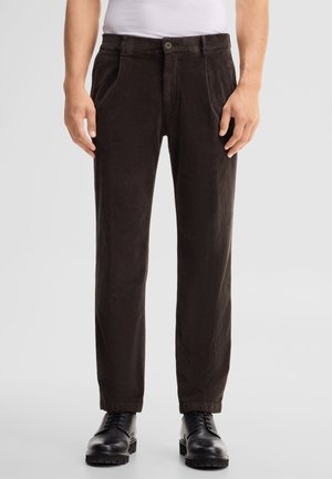 Uomo che indossa pantaloni di velluto a coste marrone scuro con pieghe, scarpe stringate in pelle nera e camicia bianca, in piedi su uno sfondo chiaro.