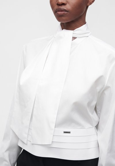 Gros plan d'une personne portant un chemisier blanc avec un détail de cravate col montant et une ceinture superposée, associé à un pantalon noir.