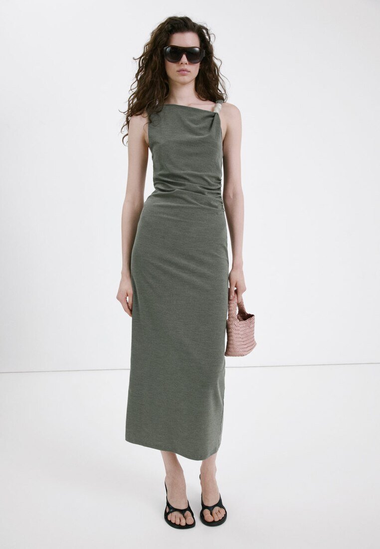 Femme portant une robe midi sans manches vert olive, de grandes lunettes de soleil noires, des sandales noires, et tenant un petit sac à main tissé rose.