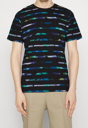 Man wearing a black short-sleeve T-shirt with horizontal blue, green, and purple stripes and beige pants, standing with arms relaxed.