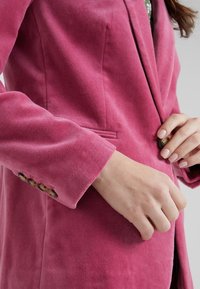 Fuchsiový blazer s matnou texturou, jednobodovým zapínáním a slim fit střihem. Obsahuje klopu s výstřihem a diskrétní boční kapsy.