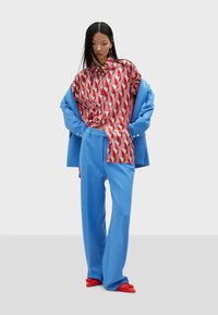 Colorful shirt with geometric pattern in reds and pinks, paired with blue wide-legged trousers and ornate red shoes. Clothing is loose-fitting.