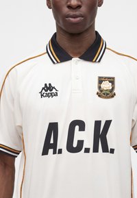 Homme portant un polo blanc avec des finitions noires et dorées, logo Kappa noir, texte A.C.K. et emblème de rose avec "Authentic 1967" sur la poitrine.