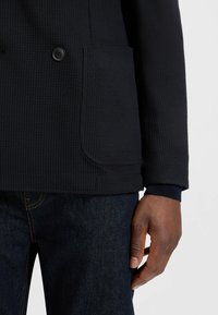 Svart strukturerad kavaj med dubbelknappar, sidoficka och marinblå stickade manschetter, i kombination med mörka jeans. Närbild av hand som håller i fickan.