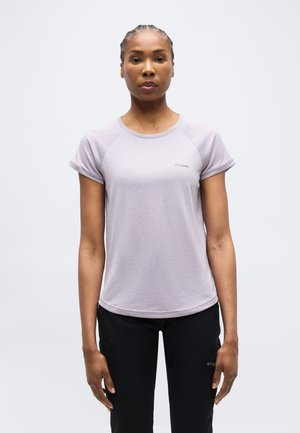 Fonott hajú nő világoslila rövid ujjú felsőt és fekete nadrágot visel, sima világos háttér előtt áll.