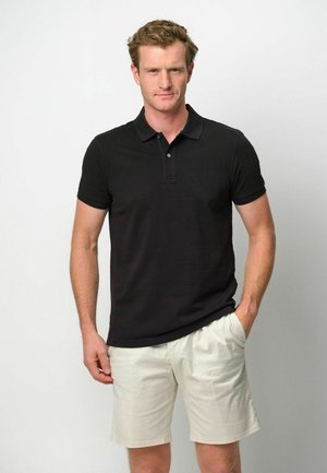 Camiseta polo negra con cuello tipo solapa, mangas cortas y tres botones. Combinada con pantalones cortos de color crema, posando frente a un fondo neutro.