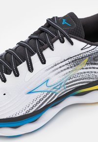 Zapatilla deportiva blanca con cordones negros y un upper texturizado. Presenta un logo azul, sutiles acentos amarillos y azules, y una suela acolchada.