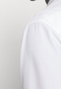 Witte overhemd met een gladde, frisse textuur. De halslijn heeft een strakke snit, met een iets zichtbare schoudernaad. Minimalistisch ontwerp.