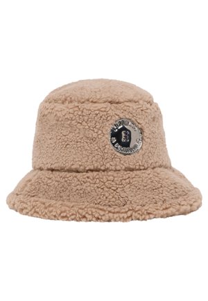 Beige sherpa bucket hat with wide brim and round black metal badge featuring text and small icon on front.