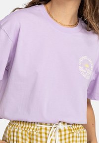 Kvinna som bär en lila T-shirt med texten "Sunshine Lovers Billabong" och en solgrafik, matchad med gula rutig byxor och ett guldhalsband.