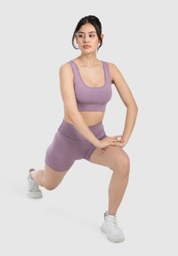 Sostén deportivo morado y pantalones cortos de tiro alto a juego, fabricados en un material suave y elástico. La modelo está realizando un estiramiento en posición de zancada.