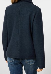 Navy blue wool jacket with a stand-up collar, featuring a textured finish and straight back design. Paired with denim jeans.