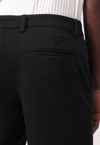 Svarte bukser laget av teksturert stoff, med et stilrent design, smale beltesløyfer og detalj med skjult baklomme.