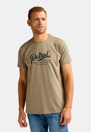 Mann mit kurzen lockigen Haaren, trägt ein hellbraunes T-Shirt mit dem "Petrol"-Logo und blaue Jeans, schaut nach links vor einem schlichten hellen Hintergrund.