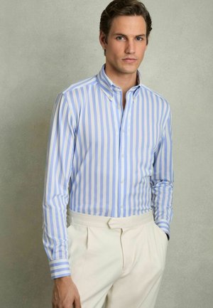 Chemise à rayures bleu clair et blanche avec col boutonné, coupe ajustée et manches longues, associée à un pantalon couleur crème.