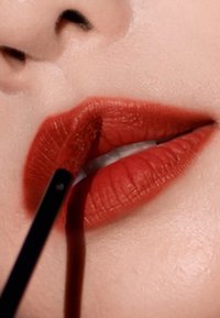 Un rouge à lèvres mat rouge est appliqué sur des lèvres pulpeuses. La texture semble lisse et crémeuse, affichant une teinte vive et riche. Un pinceau est visible en cours d'utilisation.