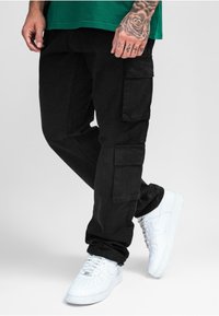 Pantalon cargo noir avec poches latérales et coupe décontractée, associé à des baskets blanches de sport. Le tissu semble robuste et lisse.