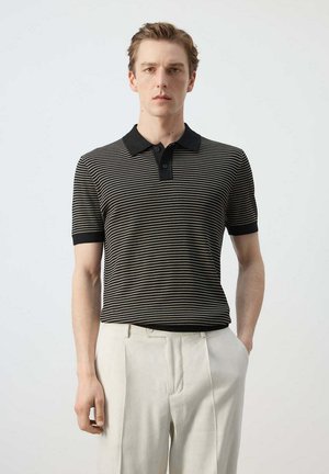 Homme portant un polo rayé noir et blanc à manches courtes et un pantalon beige clair, debout avec une main dans la poche.