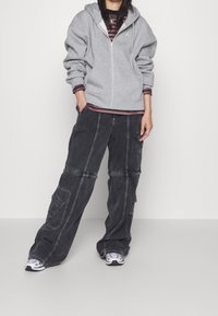 Sudadera gris con cremallera y un ajuste relajado, llevada encima de una camiseta de rayas. Pantalones cargo negros con varios bolsillos y zapatillas blancas.