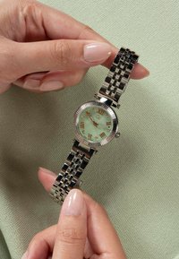 Mâini care țin un ceas de mână din metal argintiu, cu un cadran verde, cu numere romane din aur roz și marcatoare de oră din diamante, pe un fundal texturat verde.