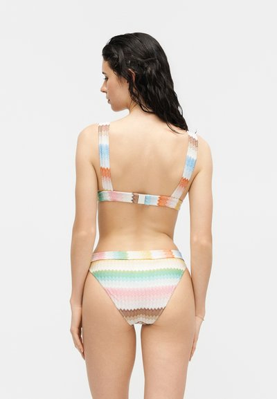 Femme de dos portant un bikini rayé multicolore avec des bretelles larges et un bas taille haute sur un fond blanc uni.