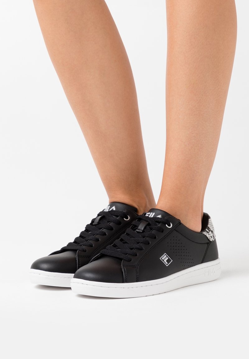 Jambes portant des baskets noires basses avec des semelles blanches et des lacets noirs, montrant un détail à motif sur la zone du talon.