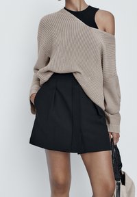 Beige neulepaita, jossa on epäsymmetrinen muotoilu, on kerrostettu mustan hihattoman paidan päälle ja yhdistetty mustiin laskostettuihin minihameisiin.