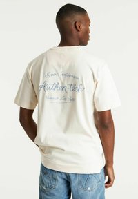 T-shirt in cotone color crema con vestibilità rilassata. Presenta un testo ricamato blu sulla schiena: "Chasin Performance Authen-tech Research Lab Ams."