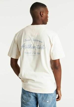 Camisa de algodón color crema con un corte relajado. Presenta texto bordado en azul en la parte posterior: "Chasin Performance Authen-tech Research Lab Ams."
