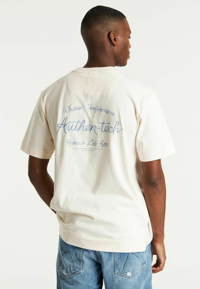 T-shirt in cotone color crema con vestibilità rilassata. Presenta un testo ricamato blu sulla schiena: "Chasin Performance Authen-tech Research Lab Ams."