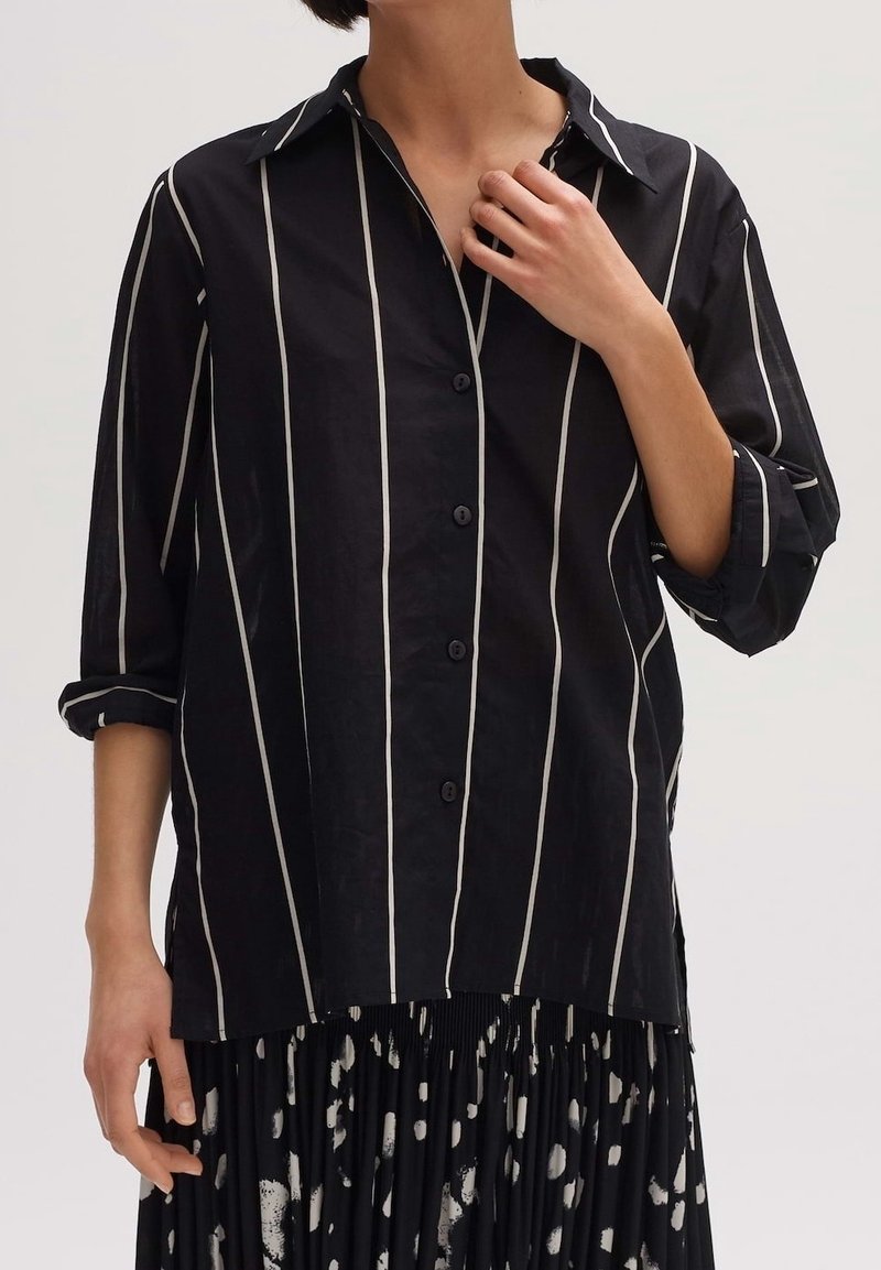 Personne portant une chemise noire boutonnée avec des rayures blanches verticales et une jupe noire avec des taches blanches abstraites, main près du col.