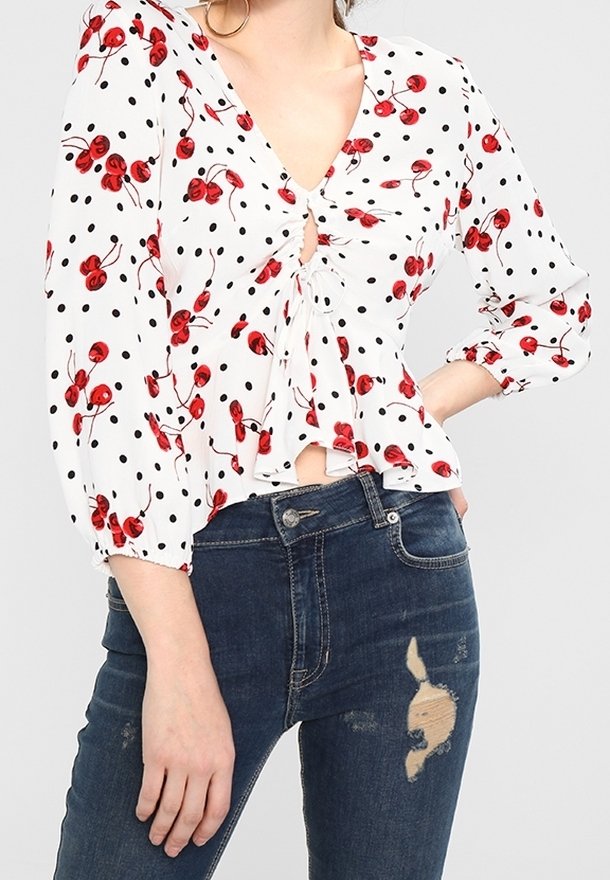 Blouse blanche à imprimé cerises rouges et pois noirs, avec un détail noué à l'avant et des manches longues bouffantes, associée à un jean bleu déchiré.