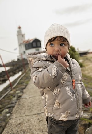 Szara kurtka puchowa w sowy, pomarańczowy suwak i ściągacze. Dziecko nosi lekki wełniany kapelusz, stojąc na kamiennej ścieżce w pobliżu latarni morskiej.
