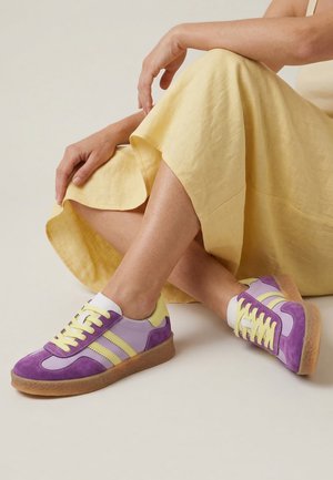 Persoon zit met gekruiste benen, draagt een gele jurk en paarse en gele sneakers met rubberen zolen op een lichte achtergrond.