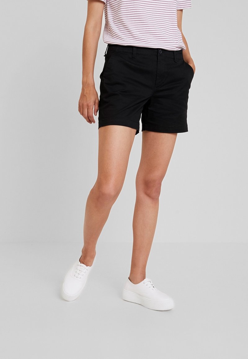 Personne portant un short noir, des baskets blanches et une chemise à manches courtes rayée rose et blanche, debout sur un sol gris.