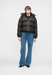 Chaqueta marrón acolchada con capucha, con cremallera frontal y puños acanalados, combinada con vaqueros de campana azules y zapatos negros de punta.