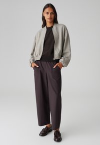 Bomber jacket grigio chiaro con polsini a coste, maglione nero, pantaloni larghi marrone scuro con tasche e mocassini neri. Texture liscia.