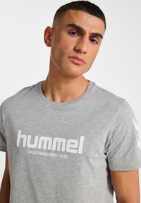 Grå bomull T-shirt med en vit logotyp "hummel" och texten "SPORTSWear SINCE 1923". Kort ärm med svarta chevron-accenter på sidorna.