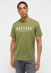 Olíva zöld pamut póló kerek nyakkivágással és rövid ujjakkal. Fehér "MUSTANG" logó található rajta, alatta további szöveggel. Egyszerű dizájn.