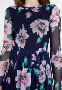 Robe fleurie avec une base marine, ornée de grandes fleurs roses et vertes. Manches longues en voile transparent, taille froncée et jupe fluide. Texture lisse.