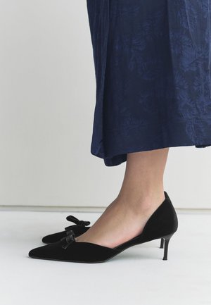 Femme portant des chaussures à petits talons noirs avec des nœuds, debout sur un sol blanc, portant une jupe texturée bleu marine.